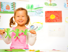 Little girl holding art project with her art on the wall behind her. Feature image.