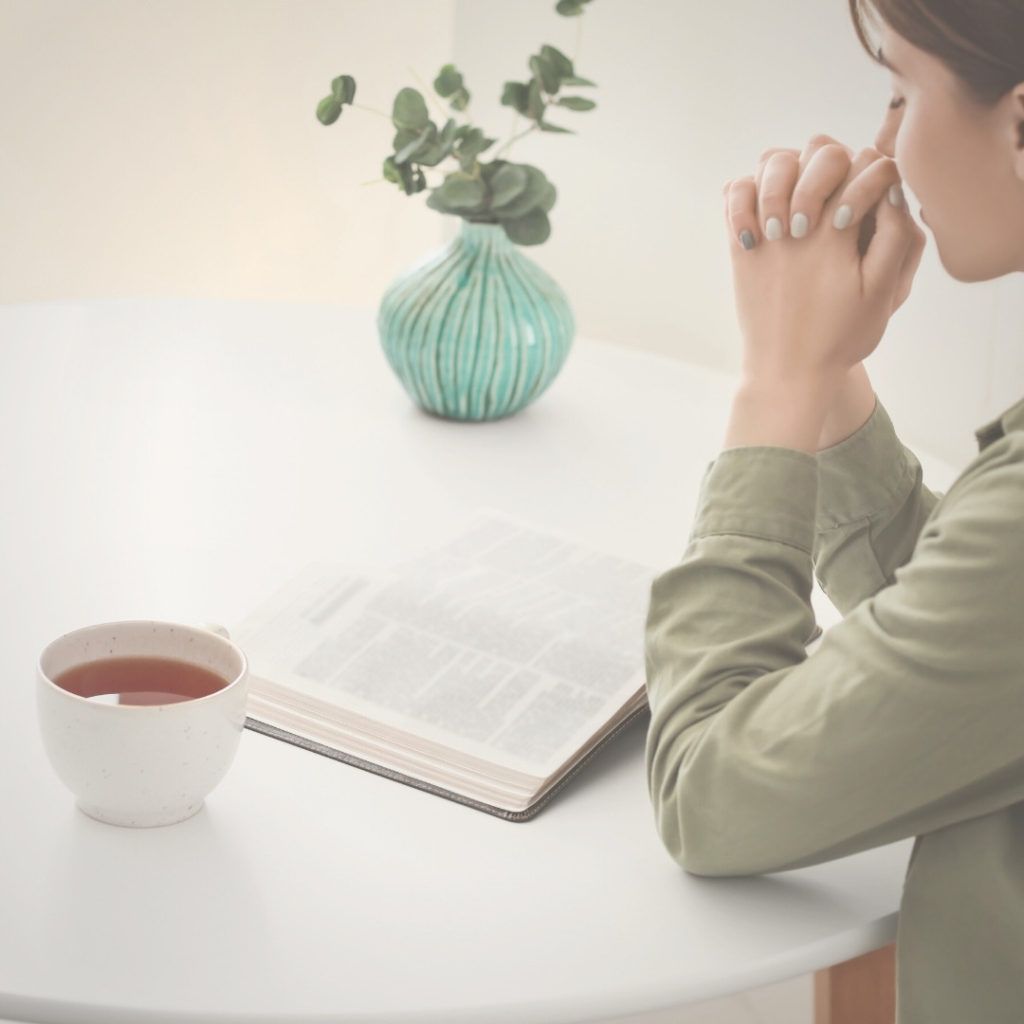Woman praying with open Bible and cup of tea on table